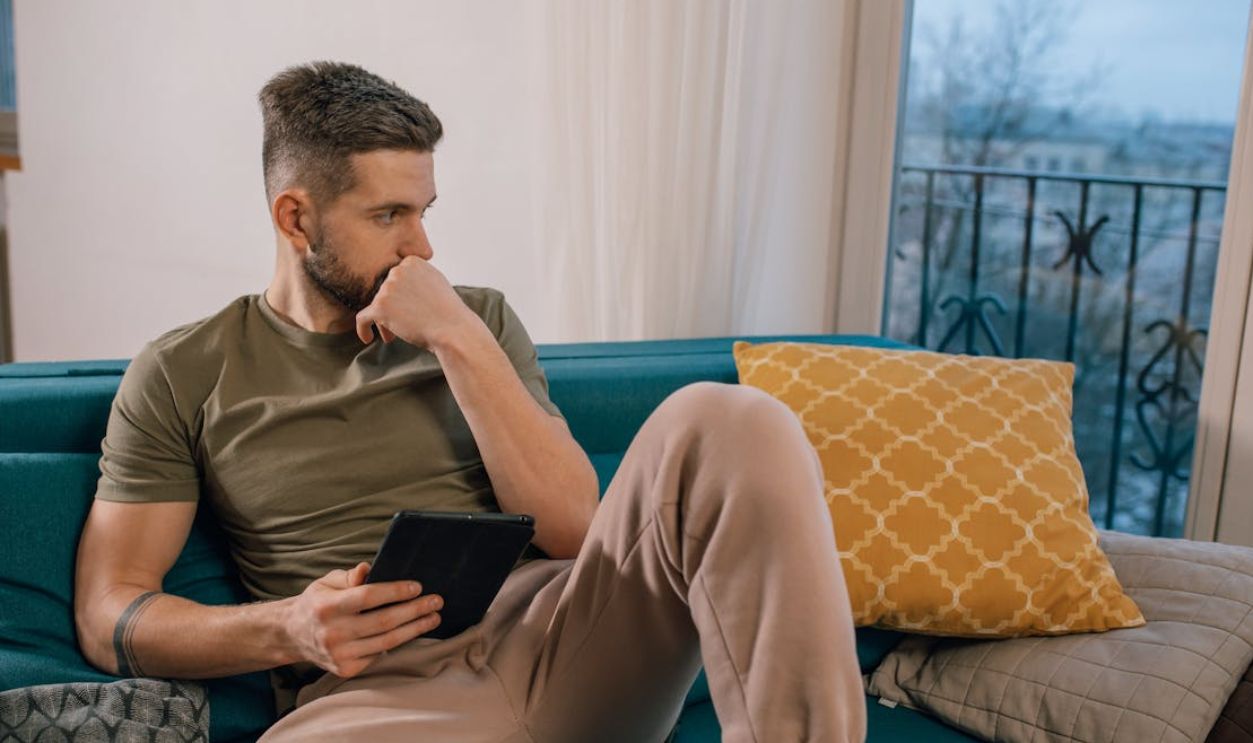 Bearded man sitting on a sofa, thoughtfully holding a tablet indoors