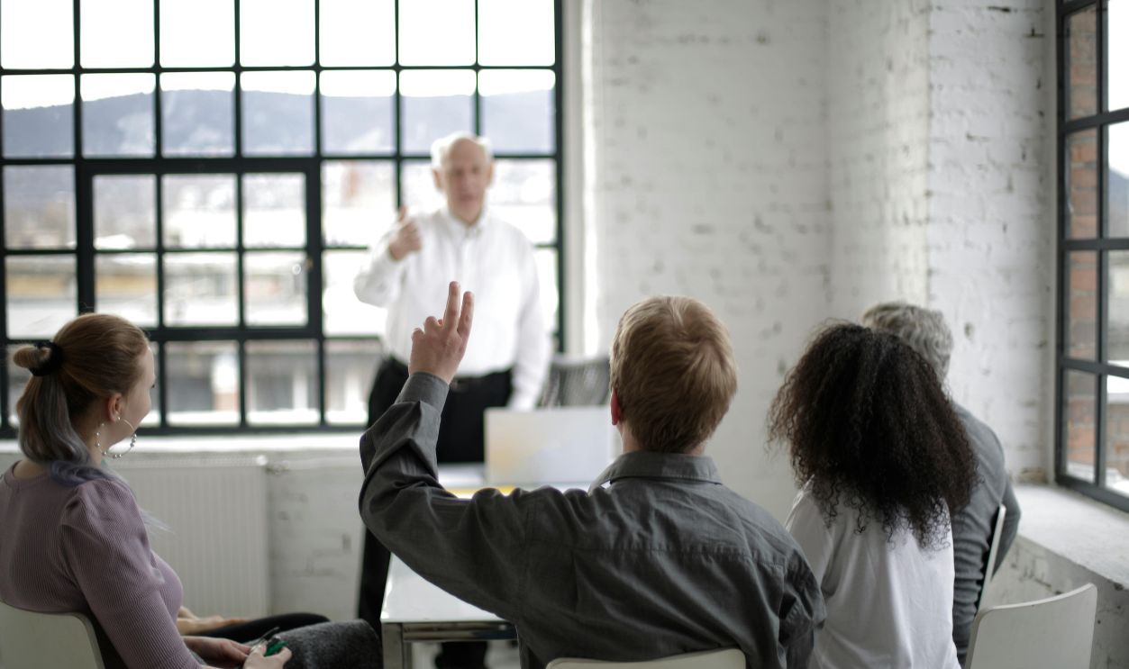Group of adults in a discussion, with one person raising a hand in a bright office space.