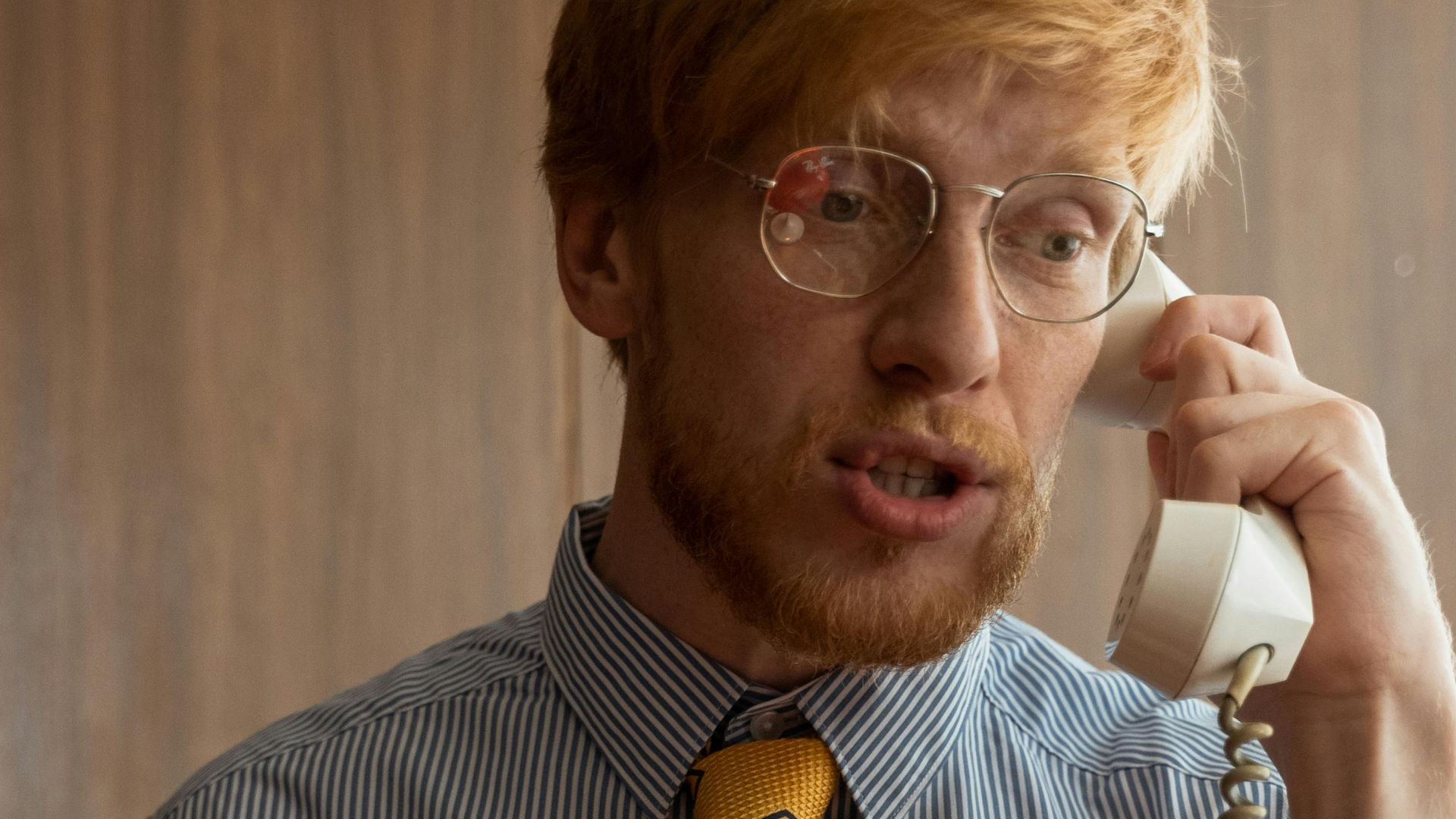 A man with red hair and eyeglasses talks on a rotary phone in a classic office setting
