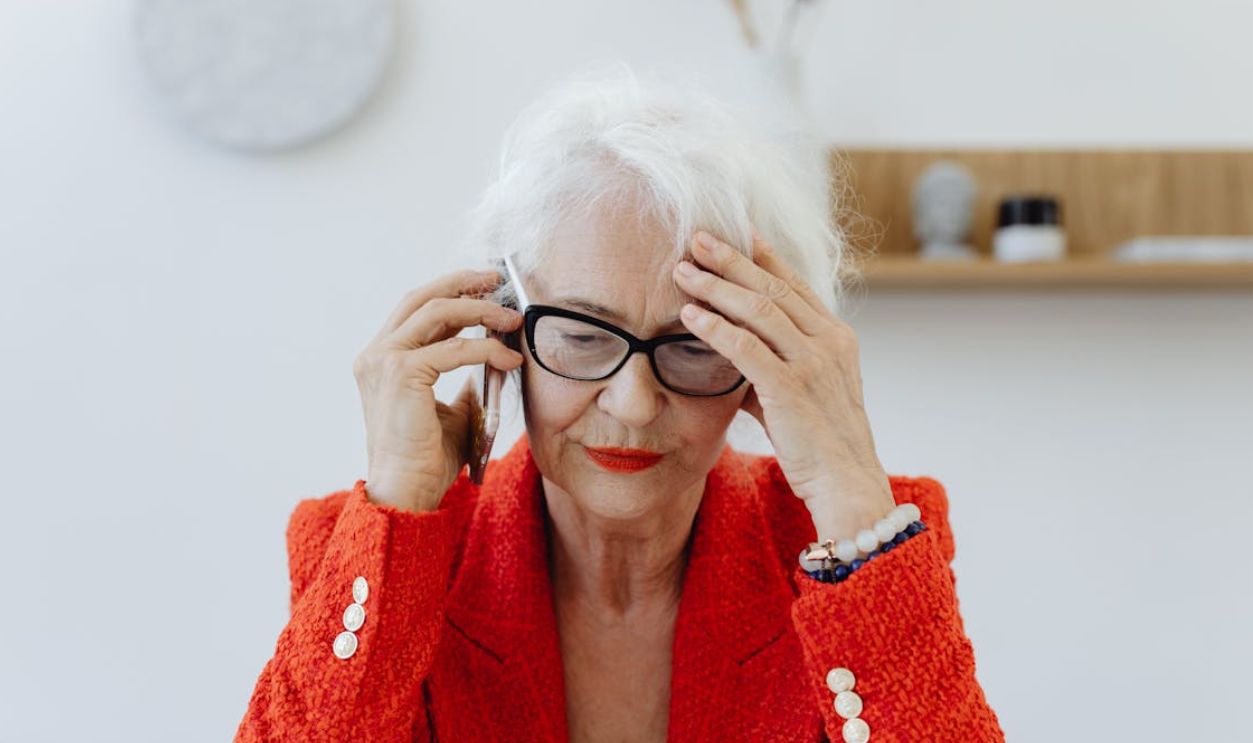 Senior woman in red blazer stressed while talking on the phone indoors.