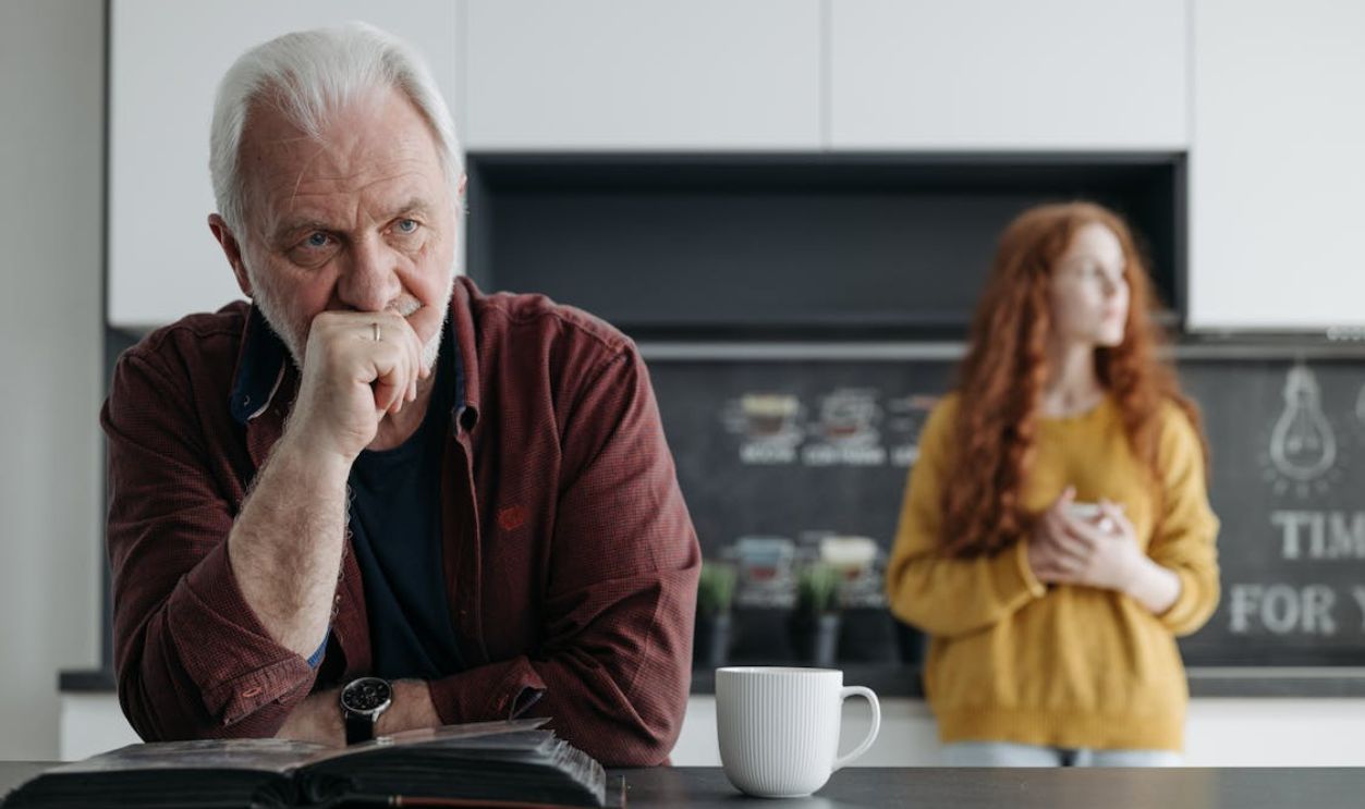 Elderly man looking thoughtful in a kitchen, with a young woman in the background, conveying a scene of contemplation and emotion.