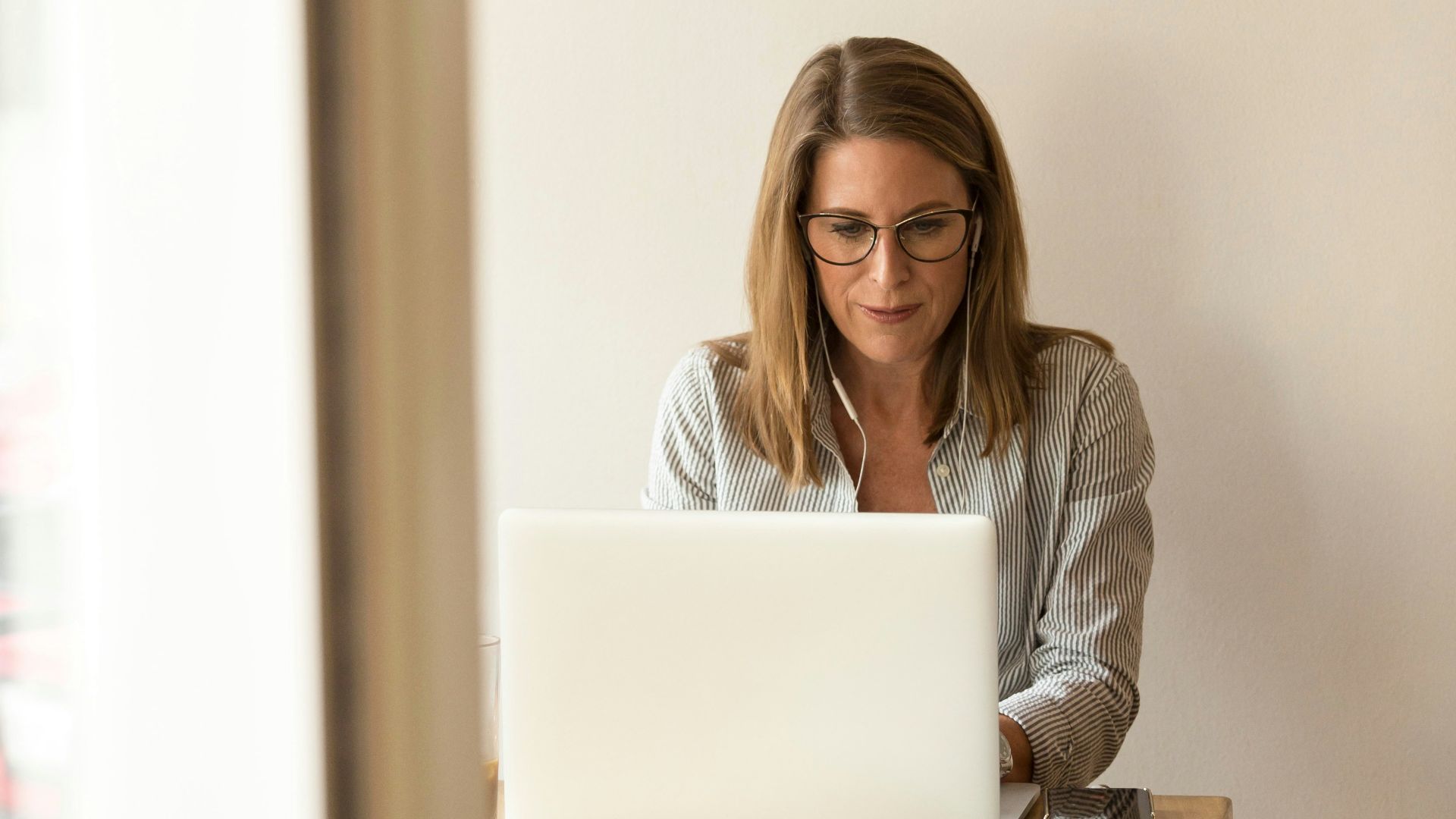 woman wearing grey striped dress shirt sitting down near brown wooden table in front of white laptop computer