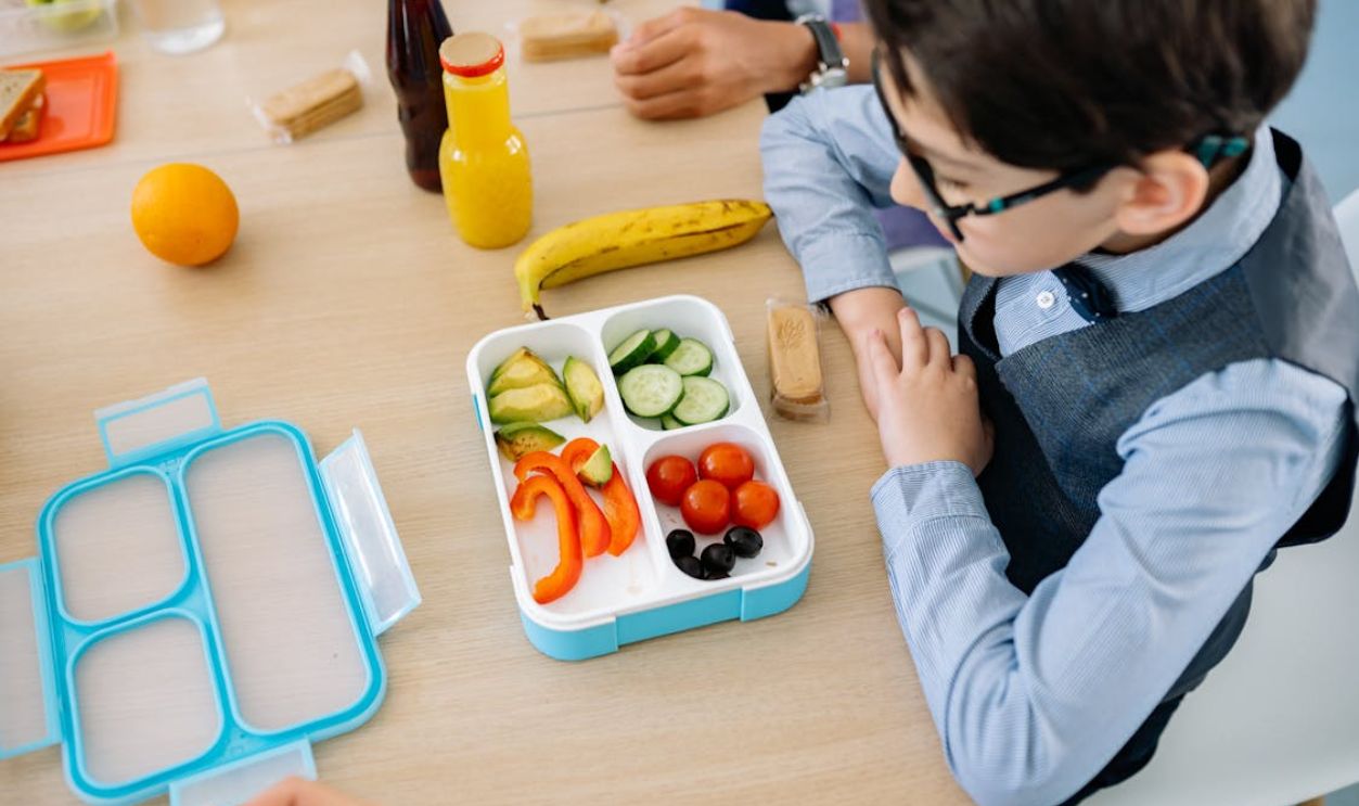 A young boy sits at a wooden table in school, enjoying a healthy lunch from a colorful lunchbox.