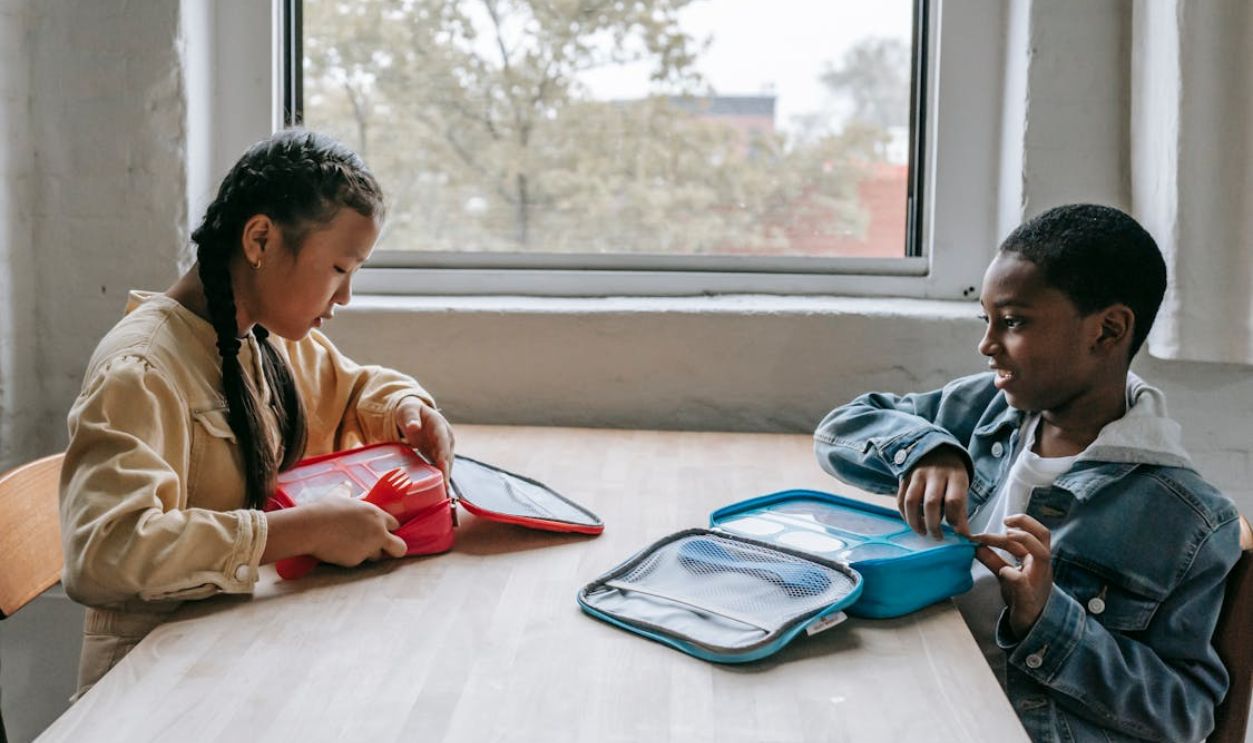 Two diverse children share lunch in a classroom setting.