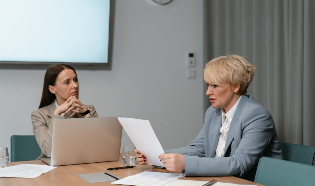 Two businesswomen in a serious meeting, discussing documents with a laptop in an office setting.