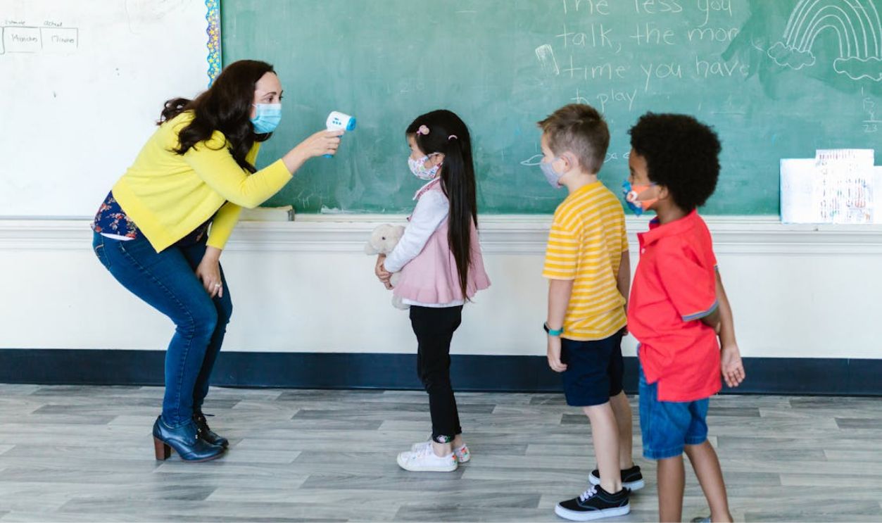 A teacher in a mask checks students' temperatures with a non-contact thermometer in a classroom setting.
