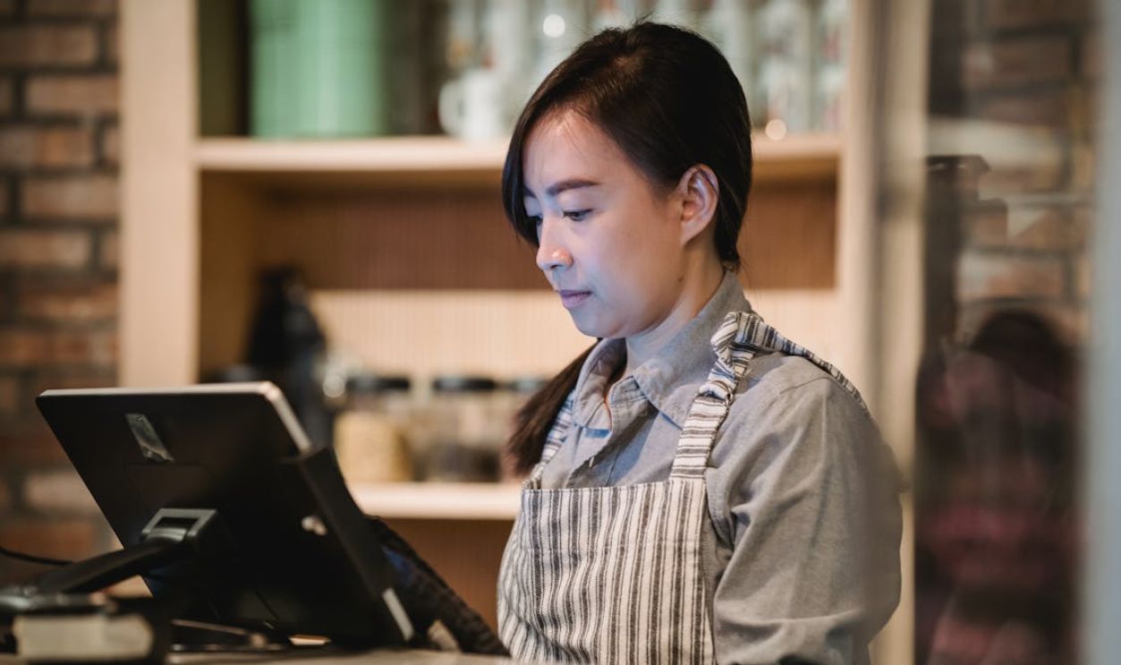 A barista in a striped apron working diligently at a cafe counter, using a point-of-sale system.