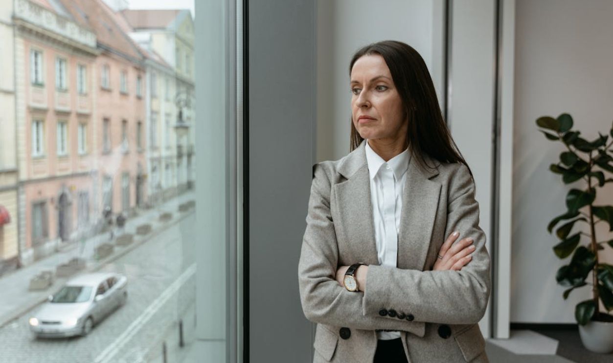 Businesswoman in grey blazer looking out office window, contemplating urban view.