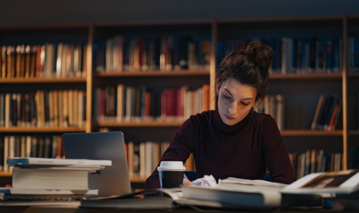 A woman sits in a library, focused on her studies with books and a laptop.