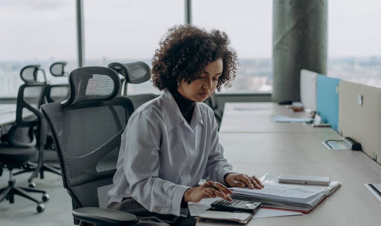 A professional woman with curly hair calculating documents in a modern office setting.