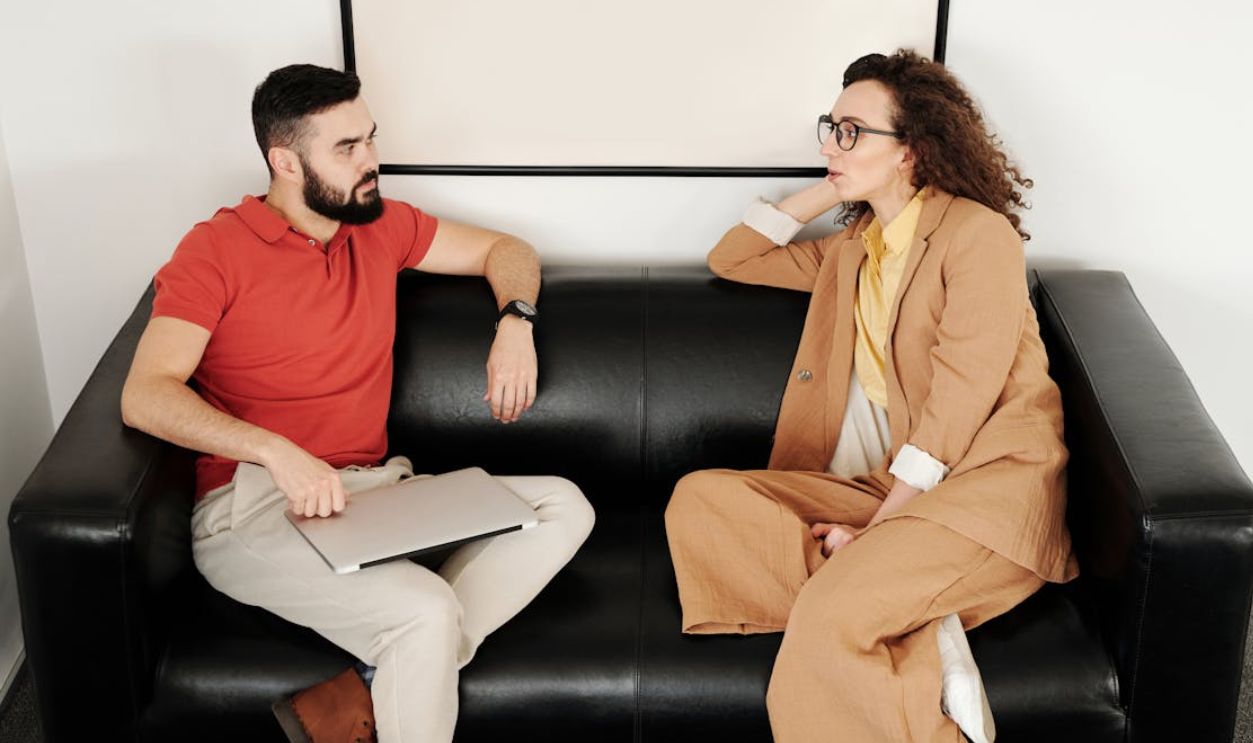 A man and woman sit on a black sofa, having a professional conversation.