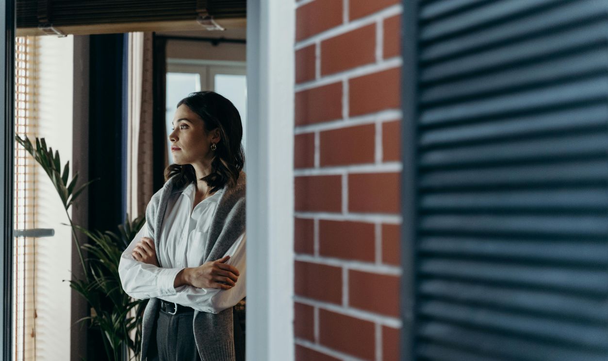 A contemplative woman stands indoors, arms crossed, gazing out the window beside a brick wall.