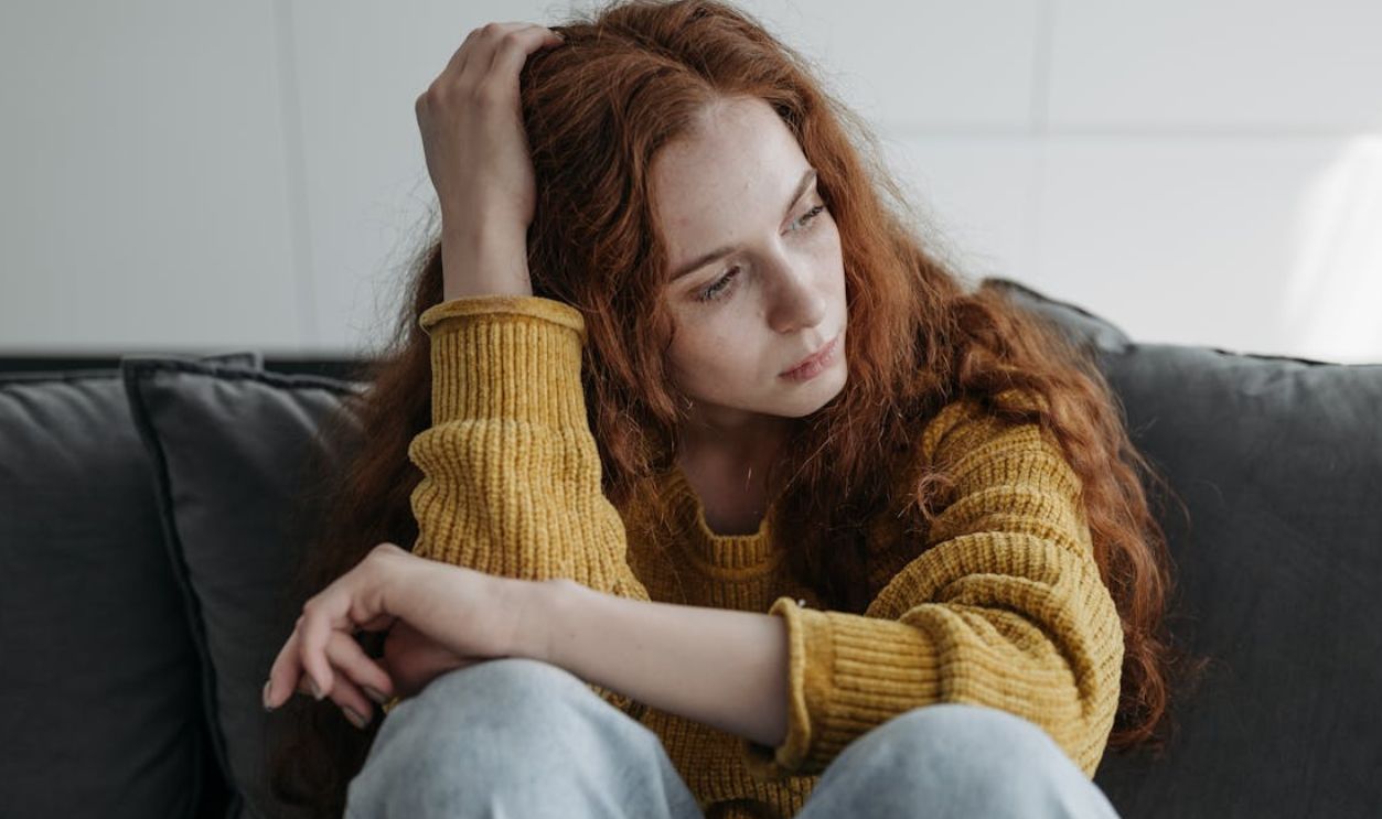 A young woman with red hair sits thoughtfully on a couch, expressing introspection indoors.