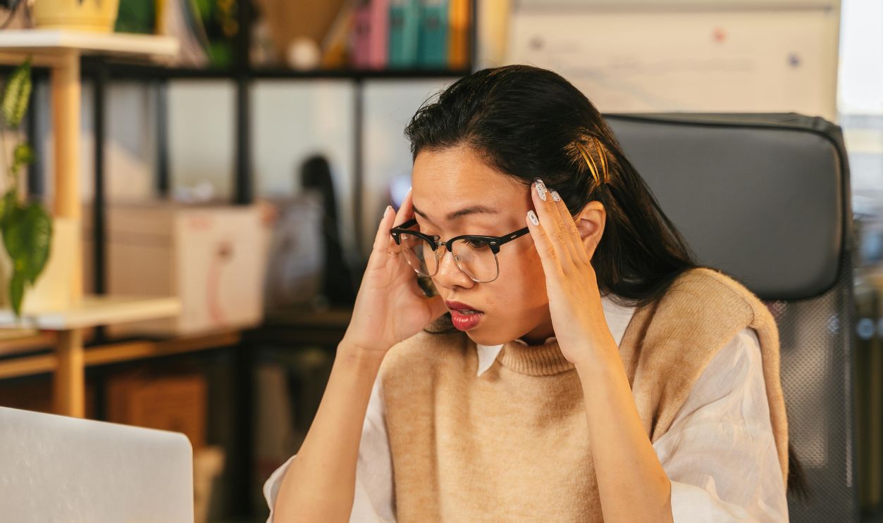 A woman with glasses shows stress while working at a laptop in an office environment