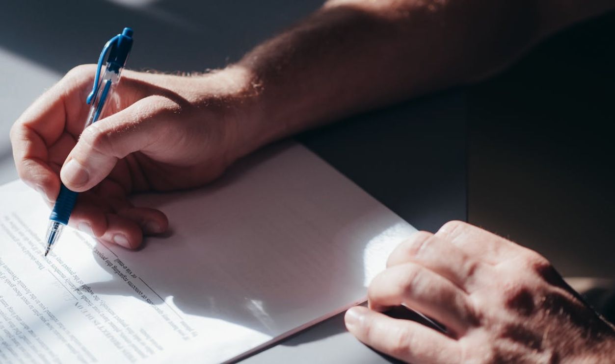 A close-up image of a person's hand signing a document with a blue pen in natural lighting