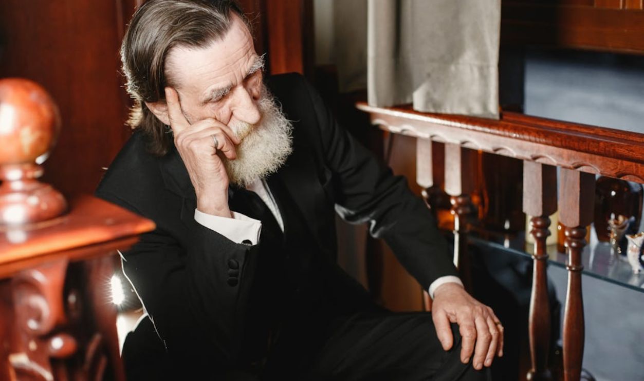 A pensive elderly man with a gray beard in a formal suit sitting indoors, captured in natural light