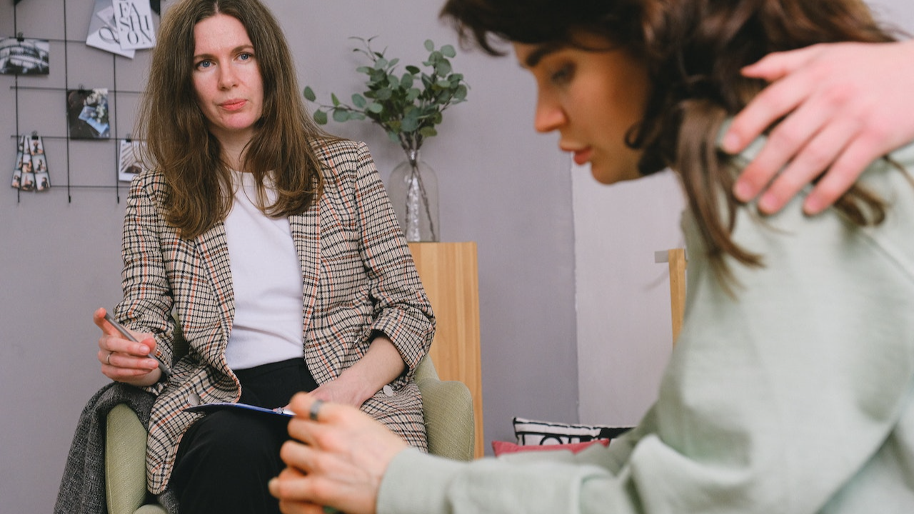 Woman with brown jacket looking at young sad woman seating on the chair and looking down.