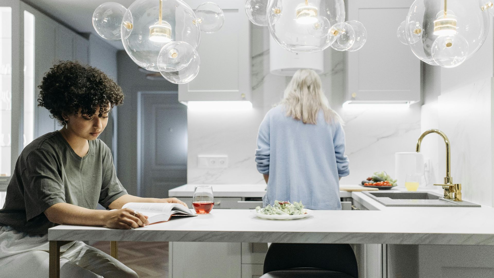 A cozy kitchen scene with a woman reading and another preparing food, emphasizing everyday life.