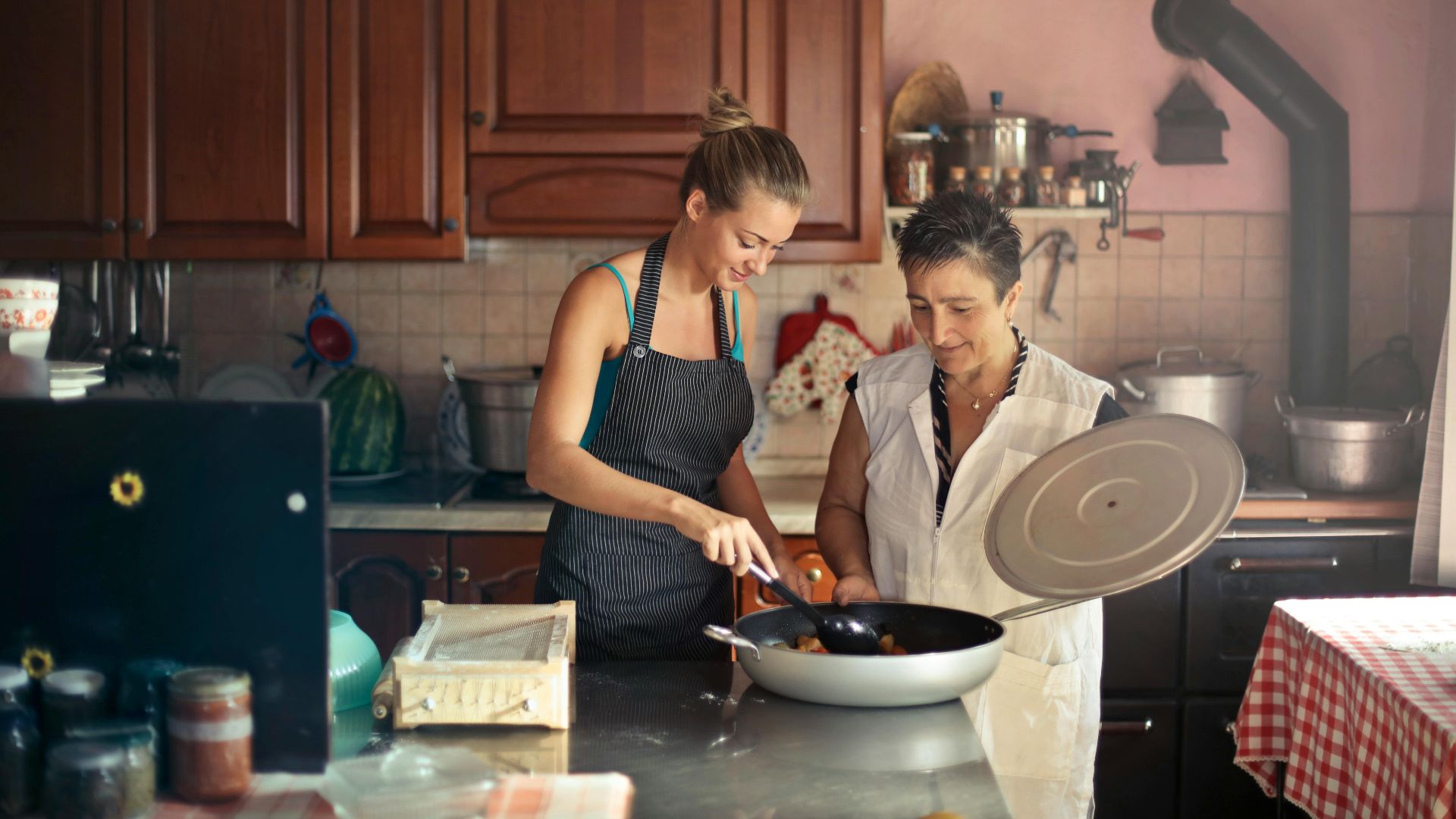 Daughter and senior mother standing at table in kitchen and stirring dish in frying pan while preparing food for dinner