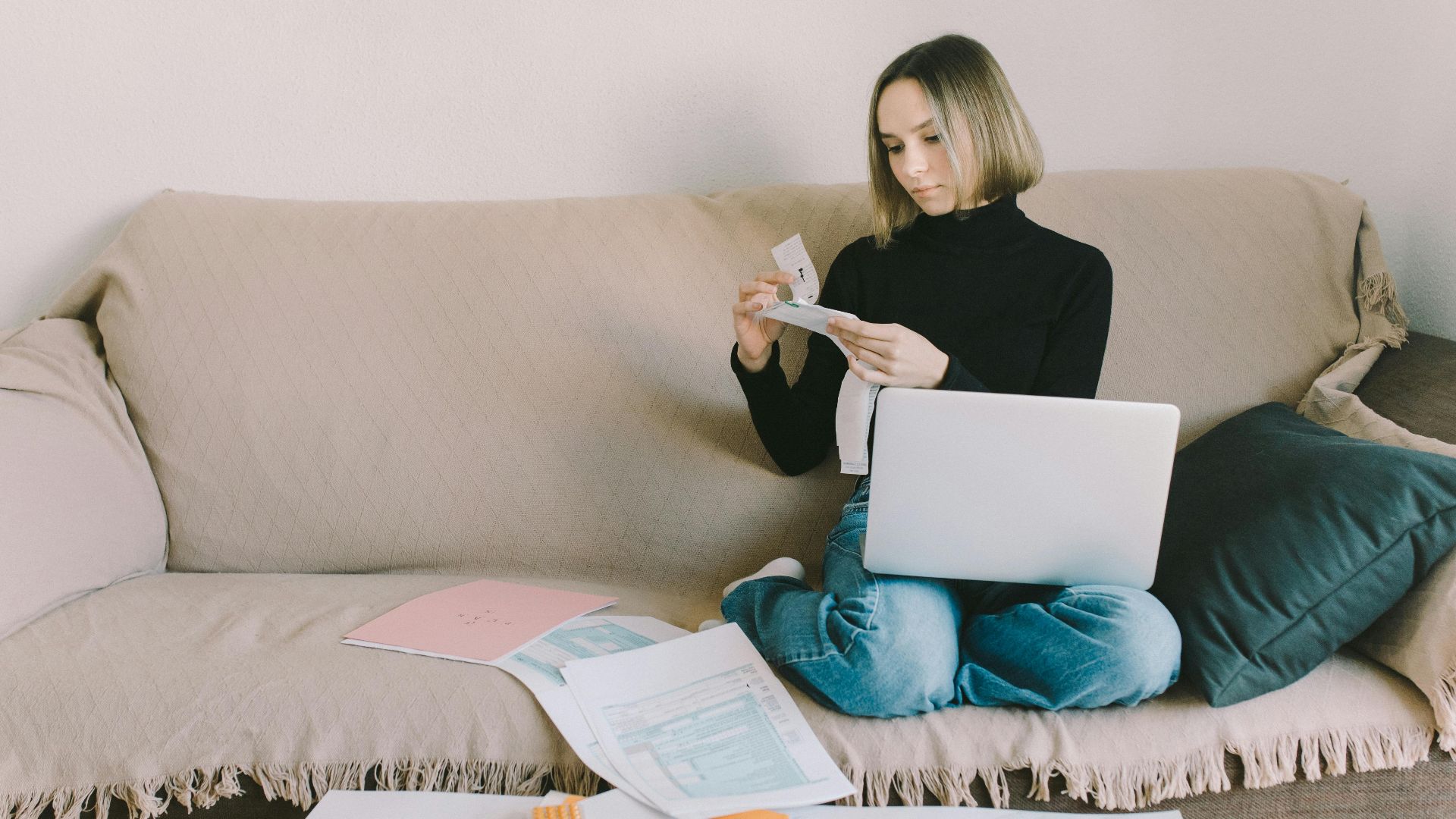A woman working from home on a sofa with a laptop and documents, managing finances.