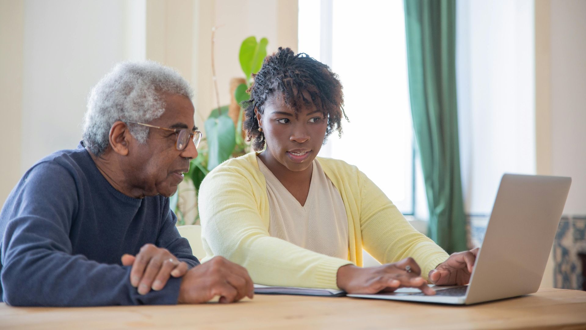 Young woman assists an elderly man with laptop indoors, highlighting intergenerational tech support.