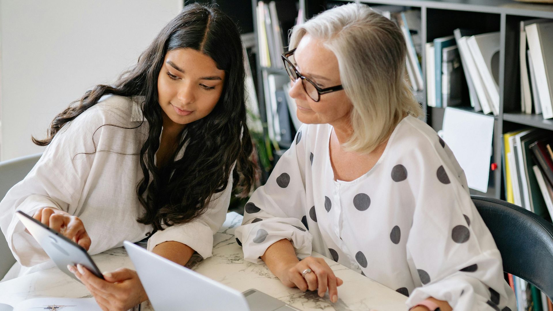 Two professional women discussing work on a laptop and tablet in a contemporary office setting.