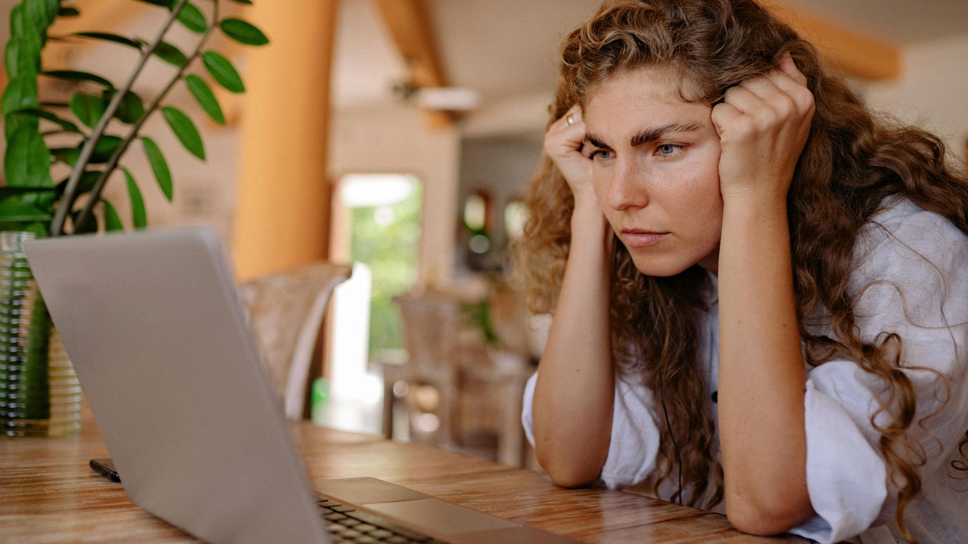 A woman with curly hair intensely focusing on her laptop indoors with plants and natural lighting.