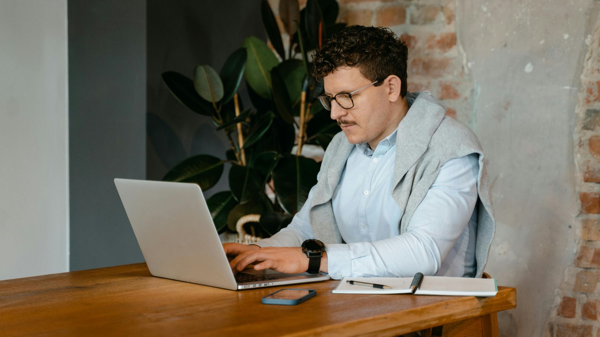 Adult man using a laptop at a wooden table in a cozy workspace with exposed brick walls.