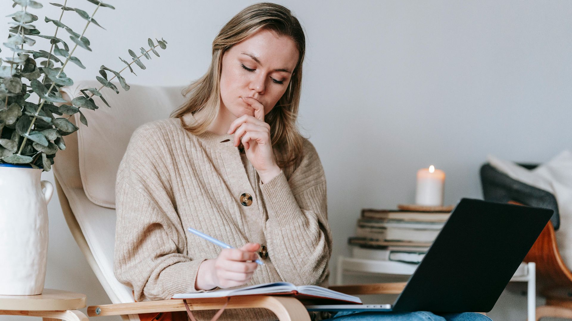 Pensive young concentrated female freelancing with netbook and diary on armchair in lounge of home