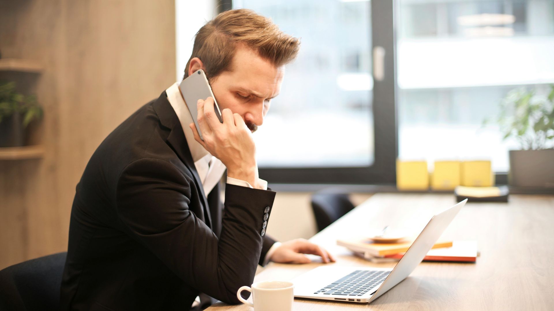 A businessman in a suit making a phone call while working on a laptop in a modern office setting.