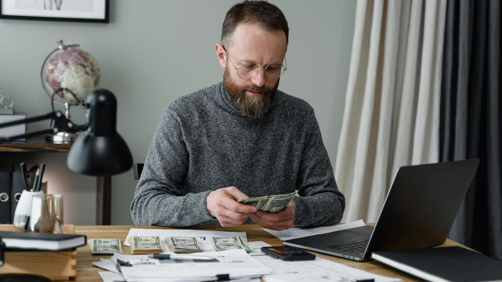 Businessman counting cash at a desk with documents and a laptop in an office.
