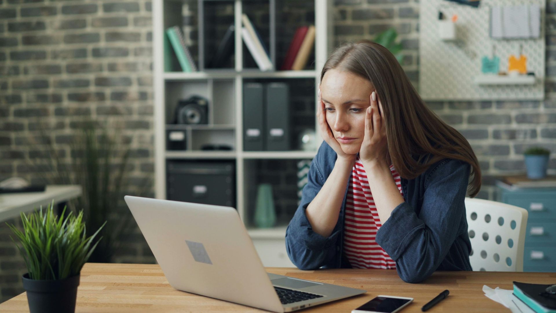 A woman appears stressed while working on a laptop in her modern office.