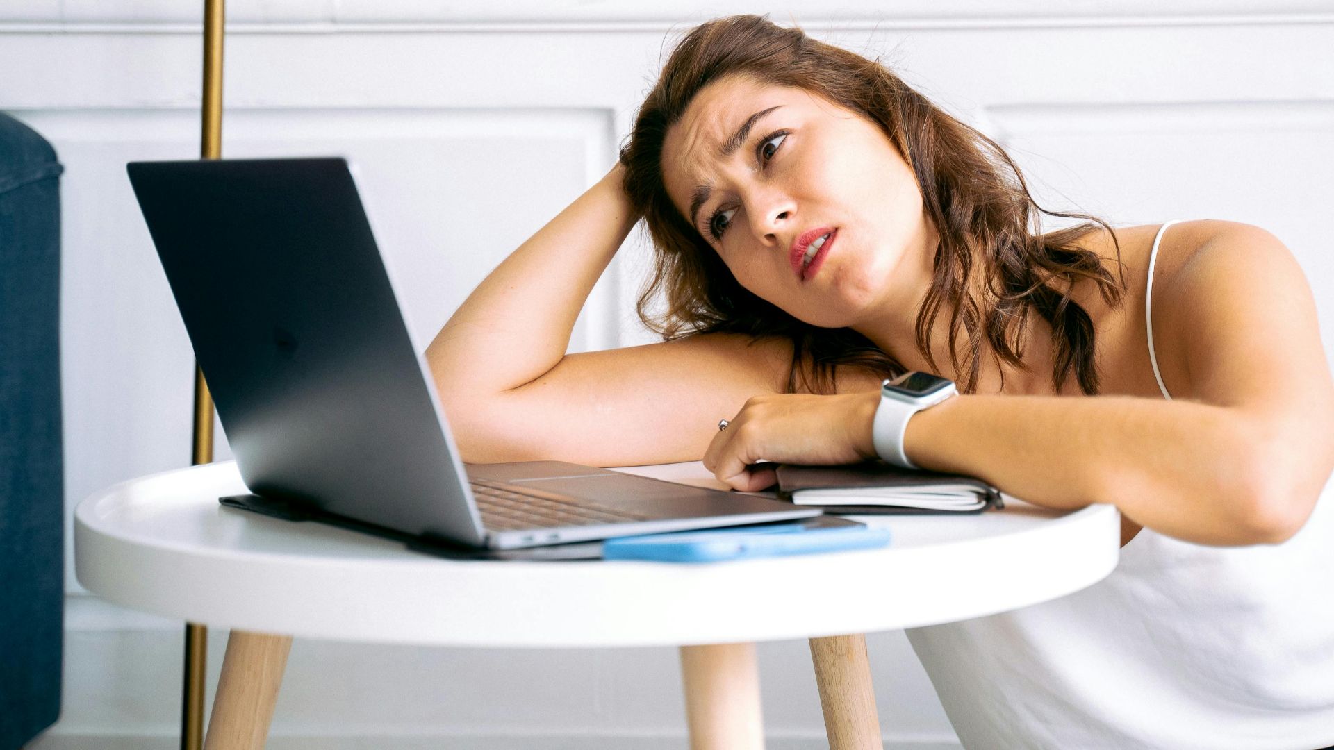 A frustrated woman sits at a desk with her hand on her head, staring at an open laptop.