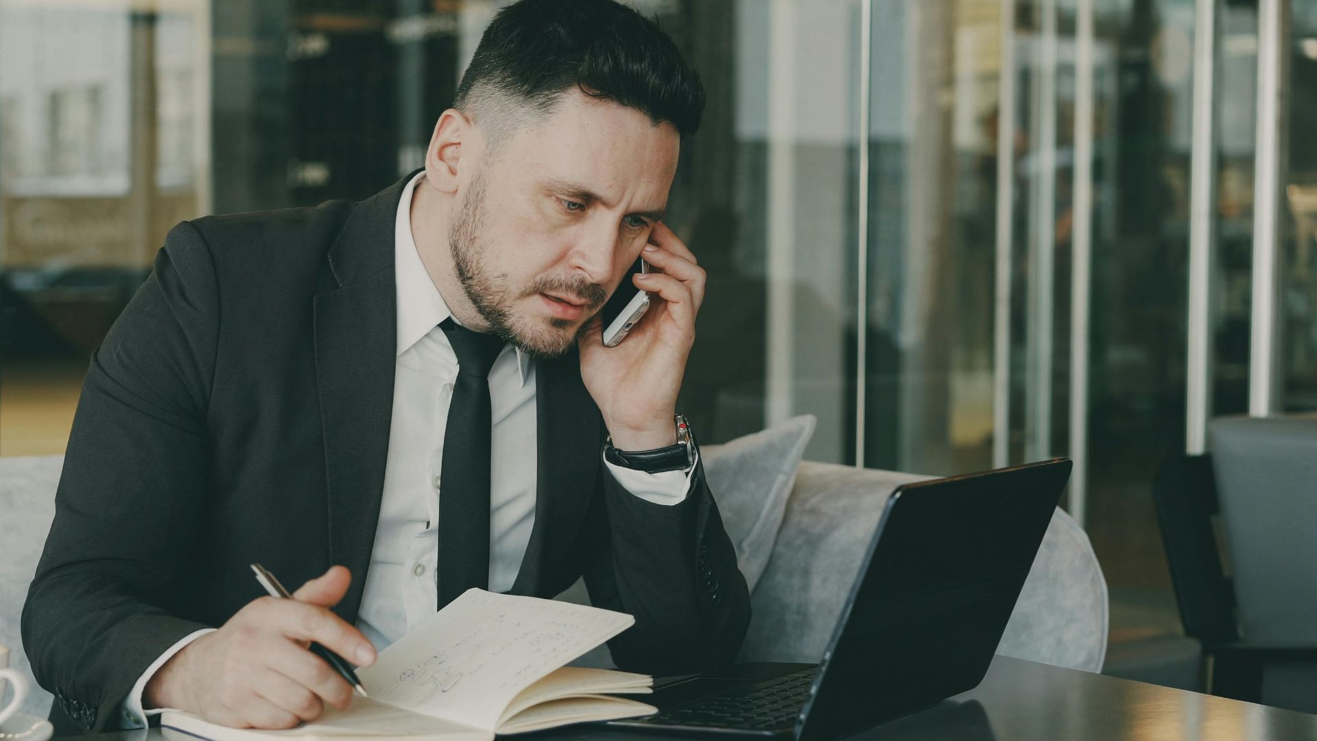 Businessman talks on phone while writing notes in a modern cafe.