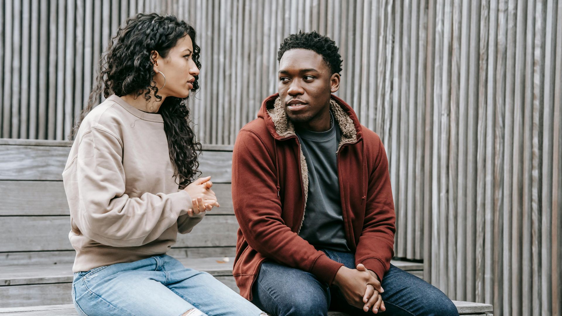 Frustrated ethnic girlfriend and African American boyfriend sitting on wooden step and looking at each other while having dispute on street
