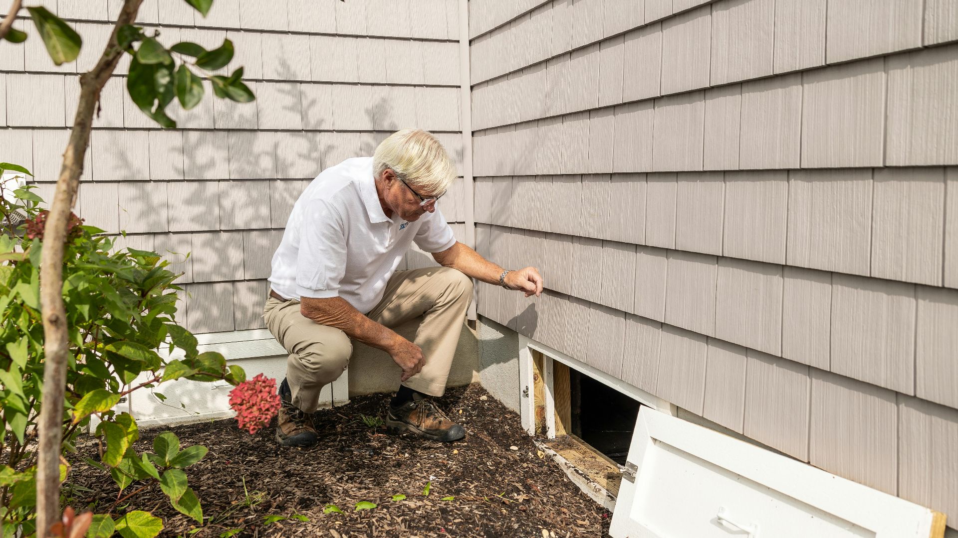 Senior adult inspecting a crawl space entrance during a home inspection on a sunny day.