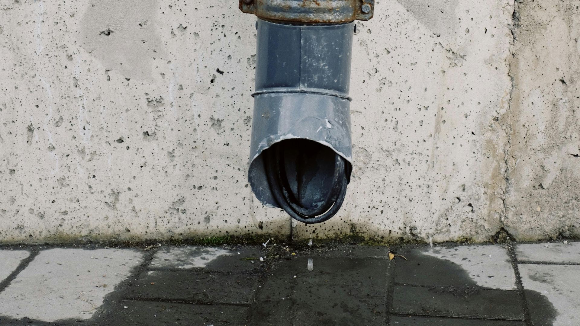 A close-up image of a metal drainage pipe on a concrete wall with visible water runoff.