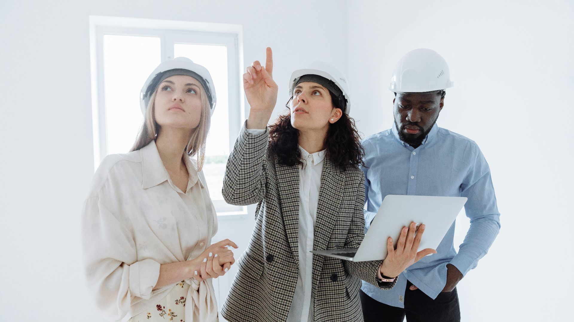 A diverse team of architects wearing helmets discusses interior plans with a laptop in a bright room.
