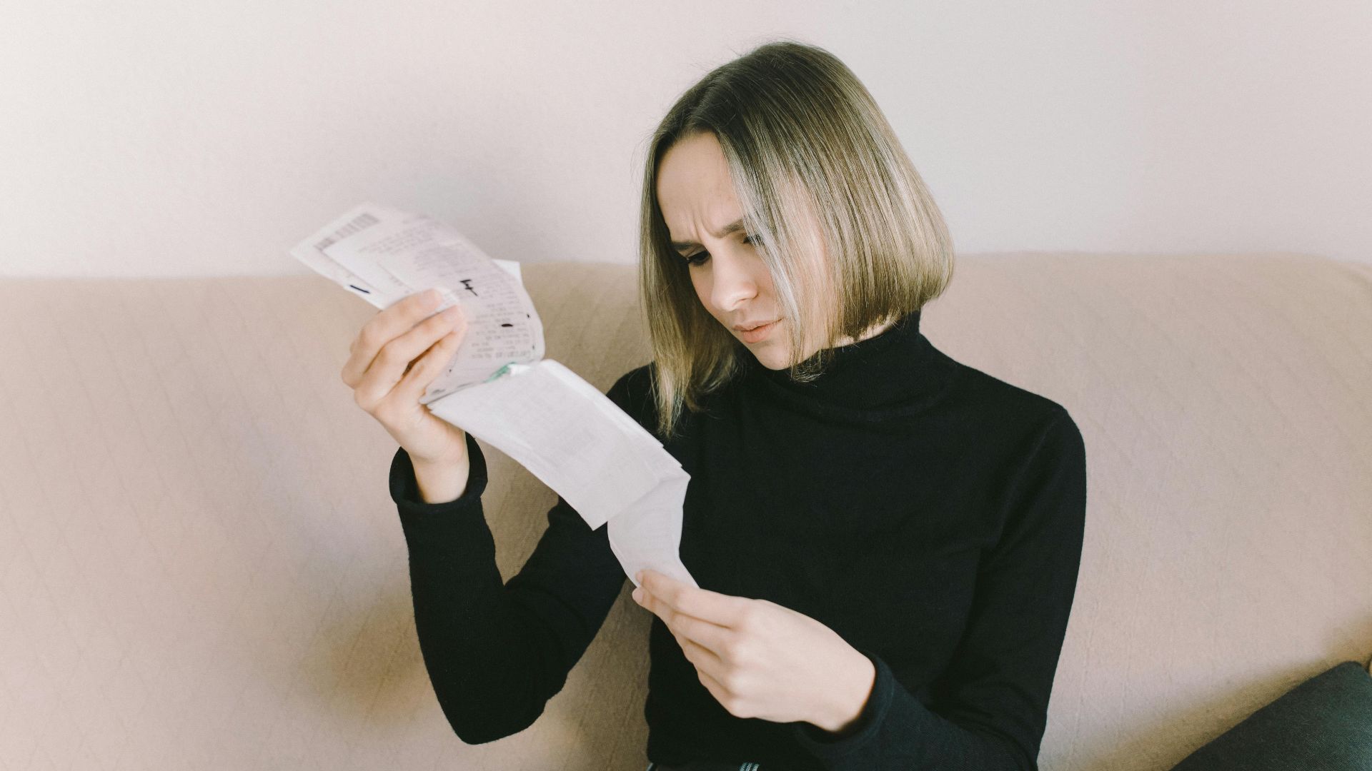 Concerned woman in black sweater examining bills on beige sofa indoors.