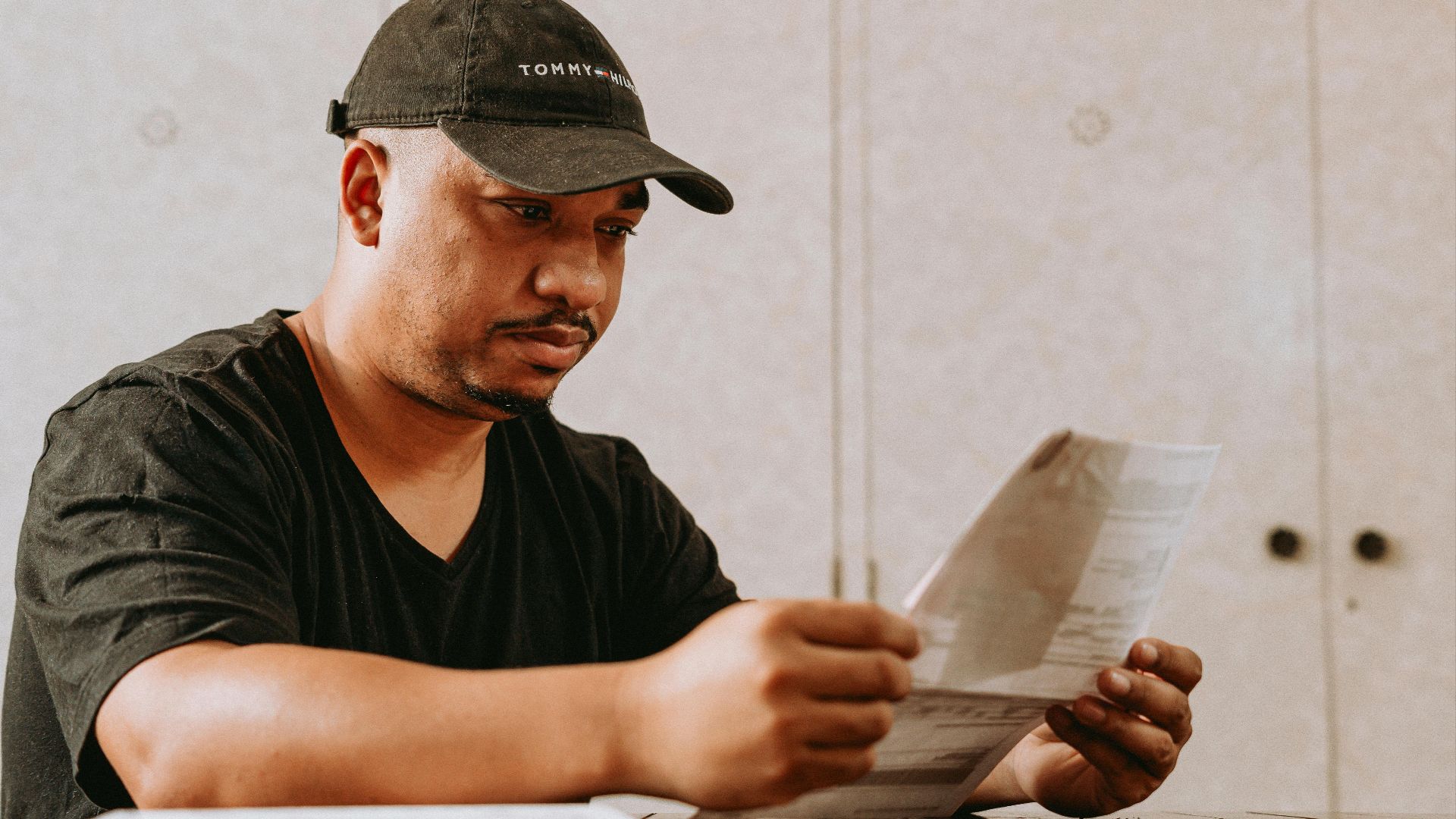 A man in a black cap reading papers at a table indoors.