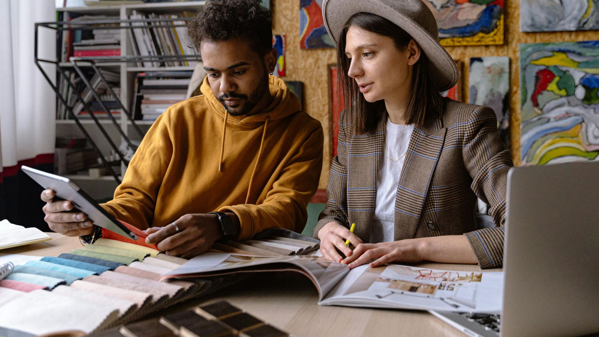 Two designers collaborate in an art-filled studio, engaging with design materials and digital devices.