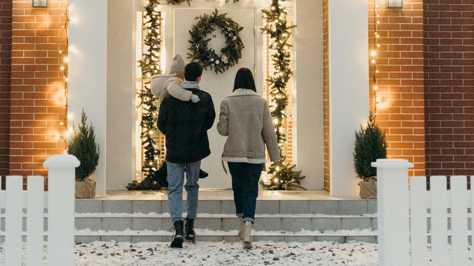 Couple approaches brick home decorated for holidays with wreath and lights.