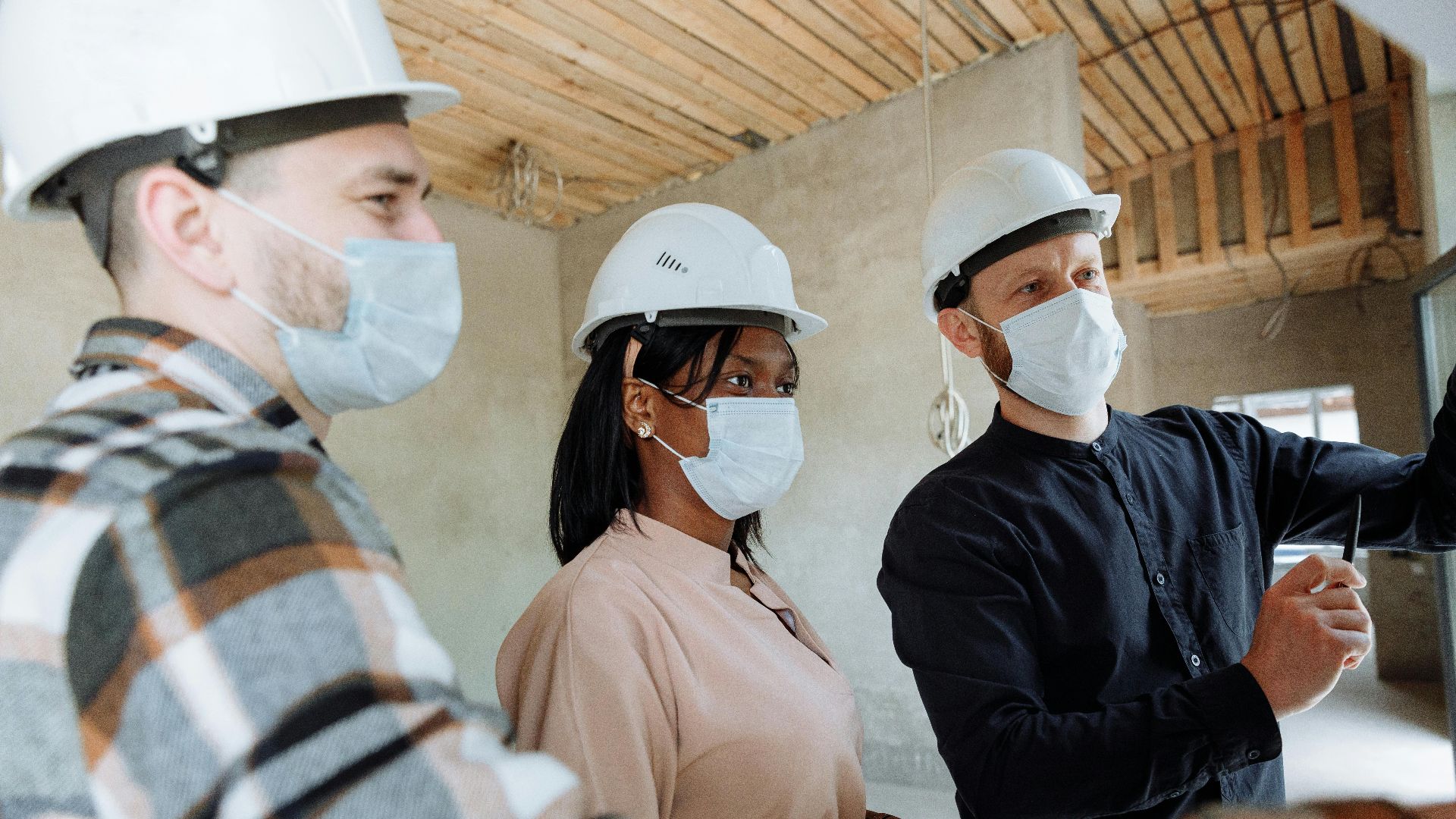 Diverse construction team wearing face masks and hard hats engaged in discussion inside a building.