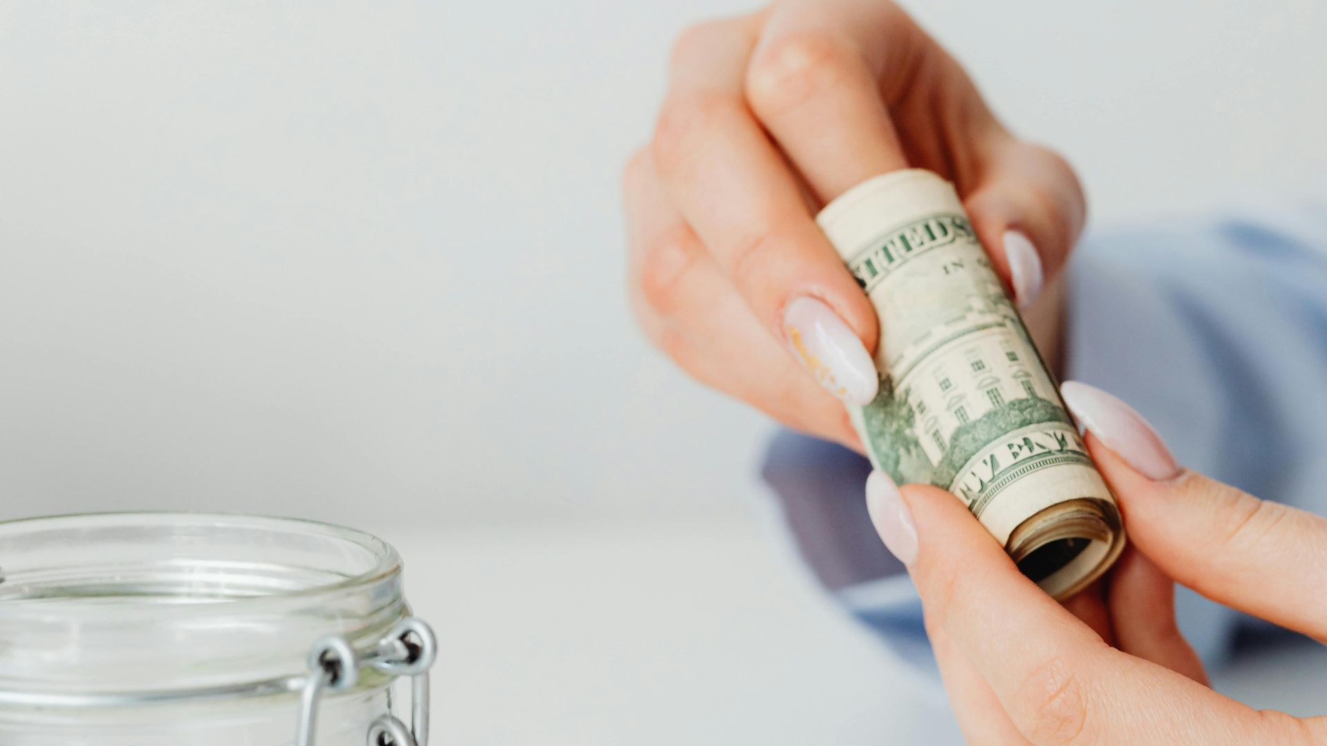 Hands holding and rolling a US dollar bill next to an empty glass jar on a white surface.