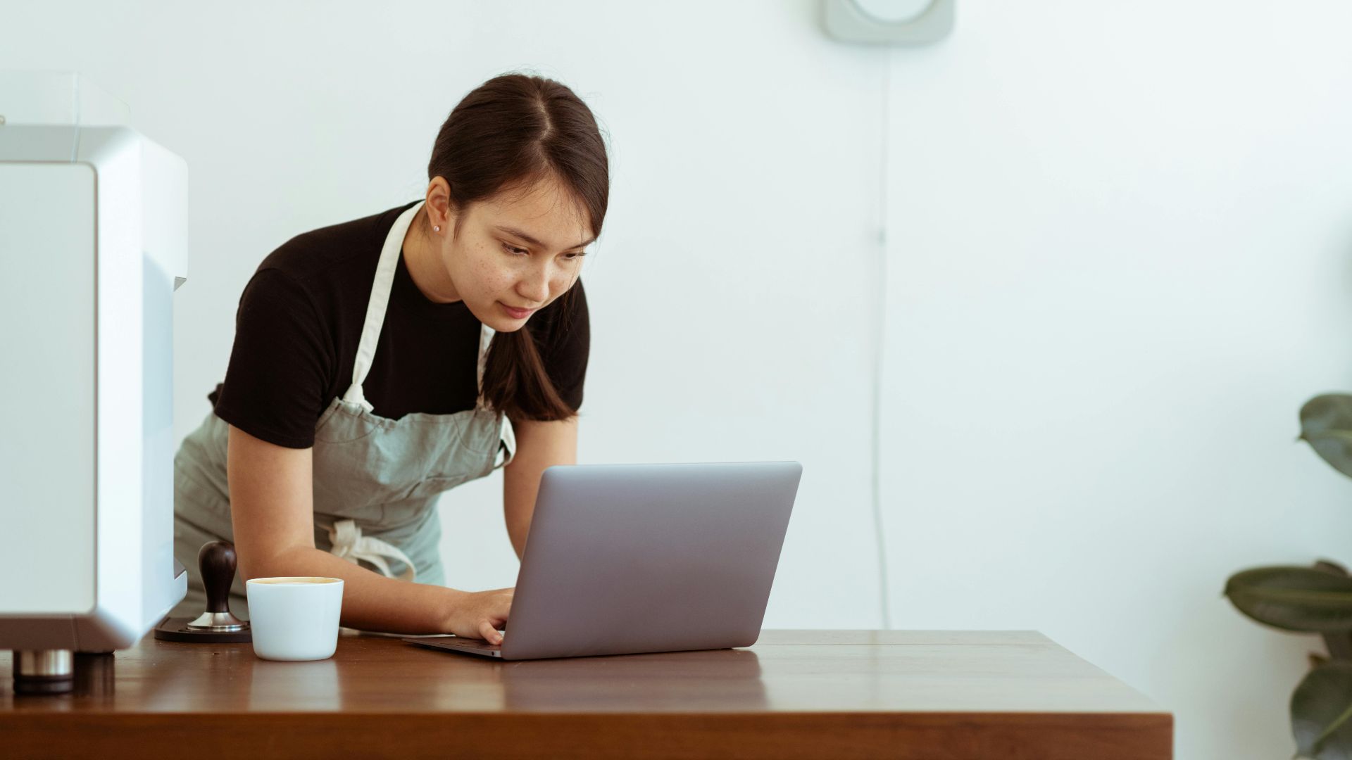 Concentrated young Asian lady in casual black shirt and apron with cup of hot drink watching cooking tutorials via modern netbook on wooden table against white wall