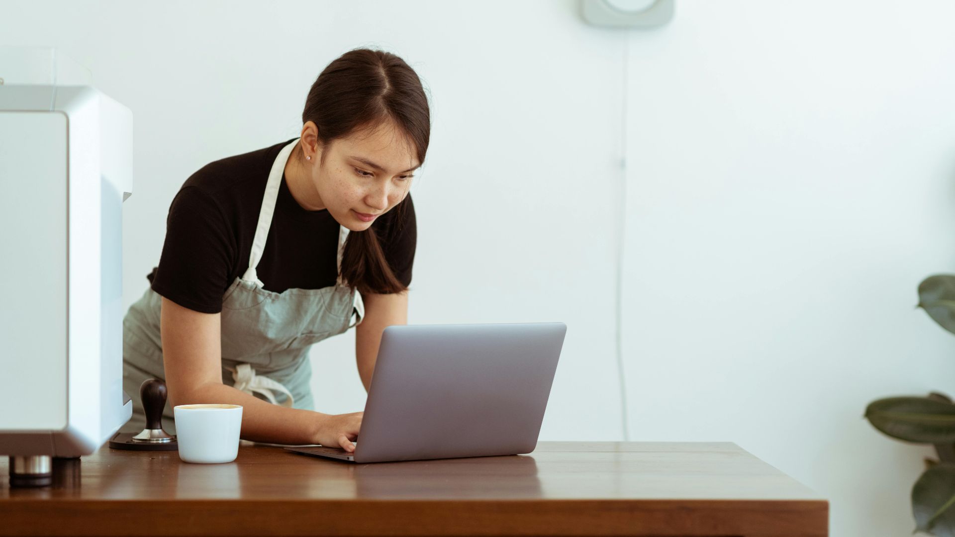 Concentrated young Asian lady in casual black shirt and apron with cup of hot drink watching cooking tutorials via modern netbook on wooden table against white wall