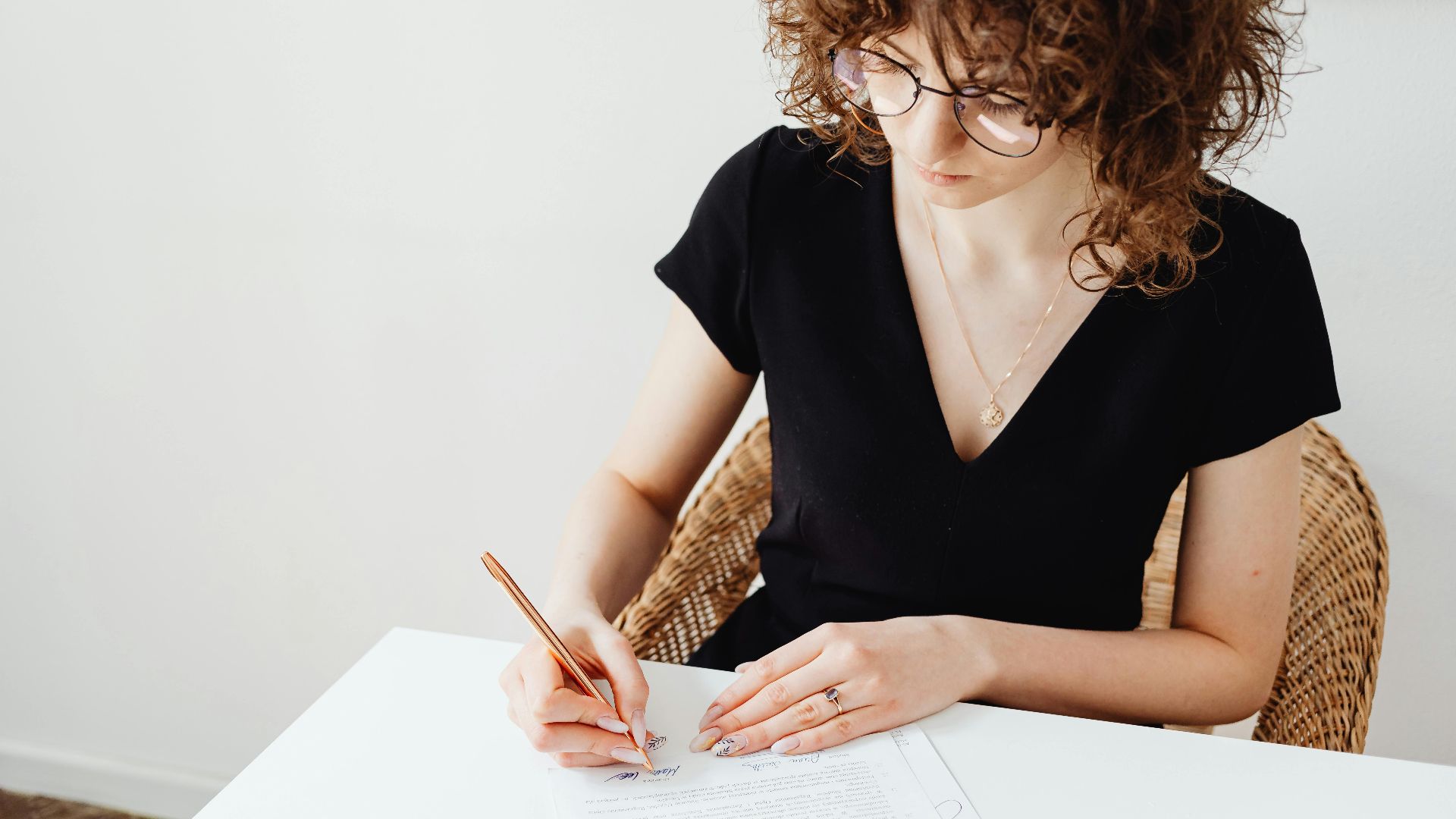 A woman in glasses and black top writing on documents at a white table, focusing intently.