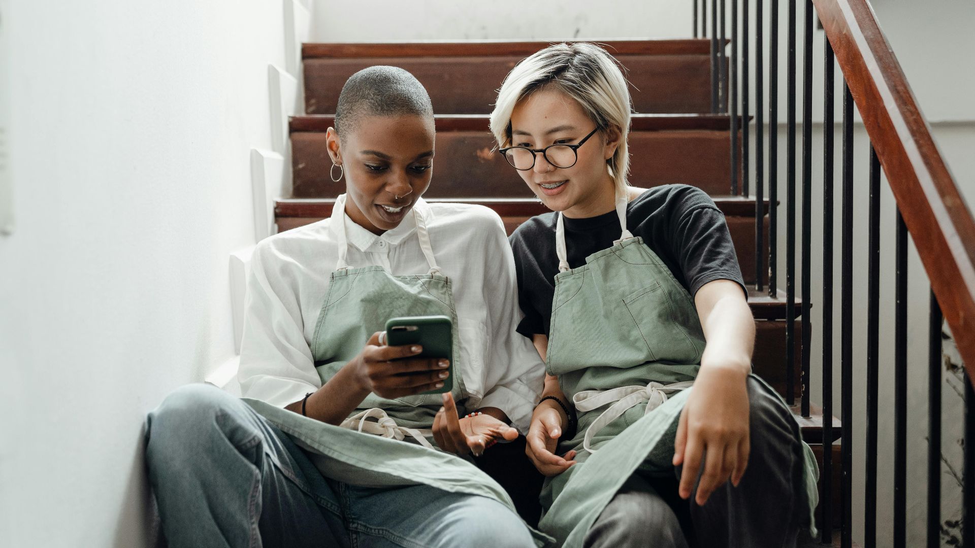 Trendy young multiracial female baristas in aprons spending time together sitting on stairs and surfing internet on smartphone