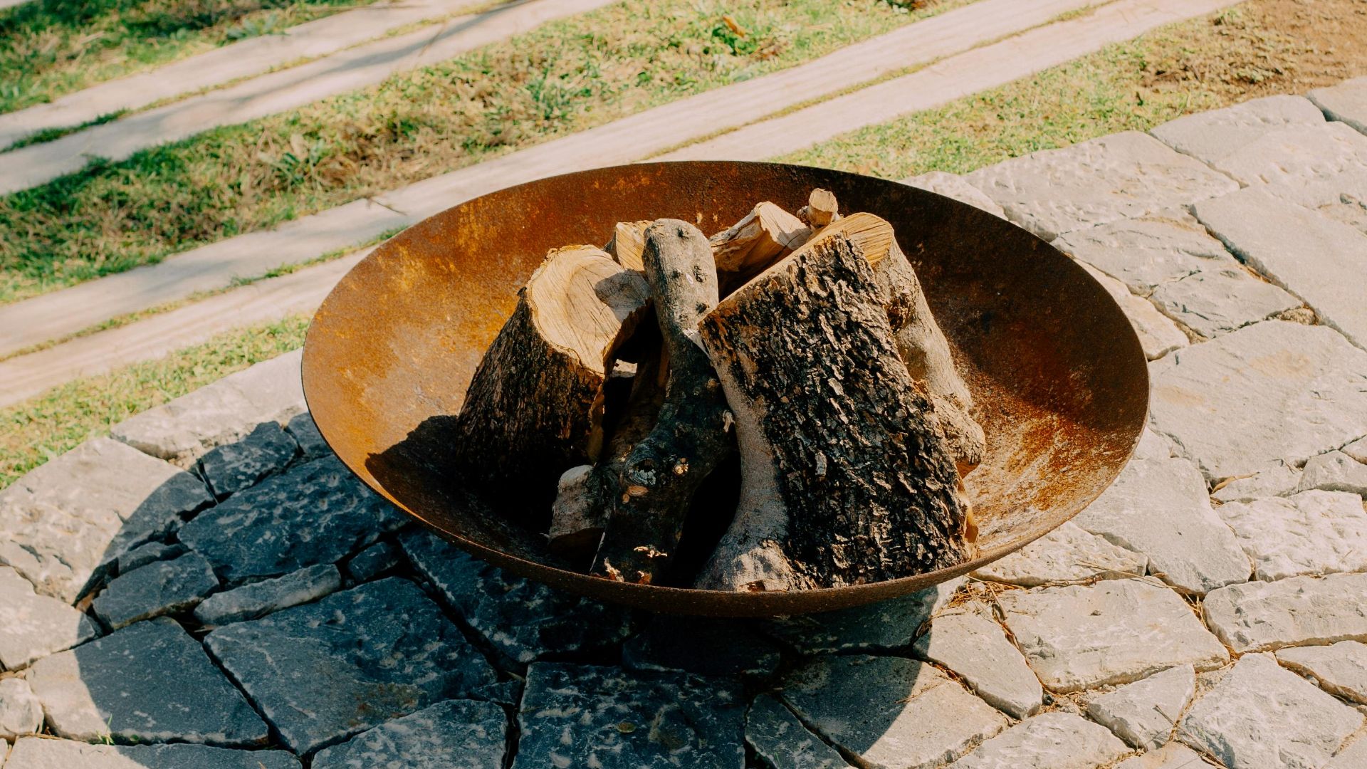 Rustic outdoor fire pit with firewood on a stone patio in a sunny Greek setting.