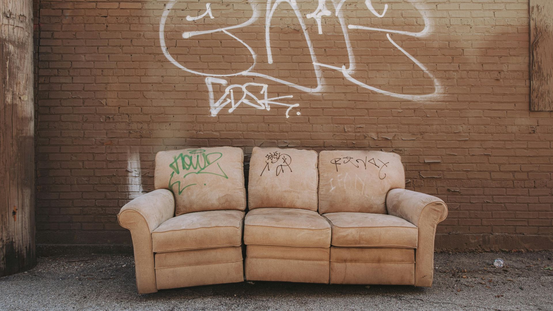 A worn-out sofa sits against a graffiti-covered brick wall in an urban alley.